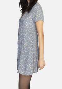 Kurzarmkleid aus hellblauem Stoff mit floralem Muster. V-Ausschnitt, entspannte Passform und knielanges Design. Kombiniert mit schwarzen Strumpfhosen.
