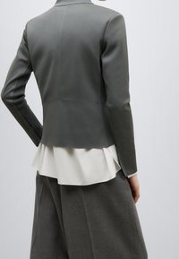 Blazer grigio aderente con colletto alto, maniche testurizzate e un volant bianco a strati all'orlo, abbinato a pantaloni grigi larghi.