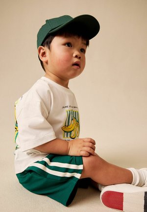 Vastsündinu, kes kannab rohelist nokamütsi, valget banaaniprindiga t-särki, rohelist lühikest pükse valgete triipudega ja valgeid tossusid, istub ja vaatab üles.