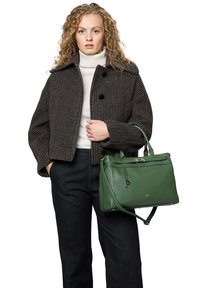 Sac à main en cuir vert avec un design structuré, poignées supérieures et une bandoulière amovible. La tenue comprend une veste courte à motif pied-de-poule et un pull.