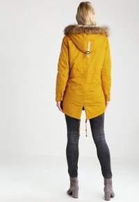 Žlutá parka s kapucí lemovanou umělou kožešinou, stahovací pas, a ventilačním otvorem na zadní straně. Nosí se s tmavými džínami a šedými kotníkovými botami. Hladký povrch.