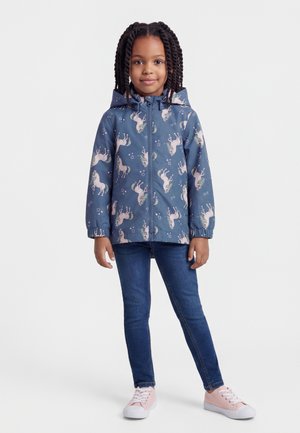 Junges Mädchen steht, trägt eine blaue Kapuzenjacke mit pinkfarbenen Einhörnern, dunkelblaue Jeans und pinke Turnschuhe vor weißem Hintergrund.