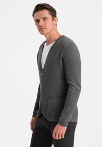 Grå cardigan med v-ringning, strukturerad stickning, träknappar och två framfickor, bärs över en vit t-shirt, tillsammans med mörka byxor.