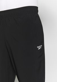 Pantalon de sport noir en tissu léger, doté d'une ceinture élastique et d'un logo Reebok blanc sur la jambe inférieure.