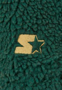 Grønn, lodden stoff med en teksturert overflate har et gullbrodert logo med en stjerne og et stilisert bokstav, som skaper en særegen aksent.