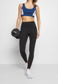 Zwarte legging in combinatie met een blauw-witte sportbh. Het model houdt een zwarte dumbbell vast en draagt witte sportschoenen. Glad, aansluitend materiaal.