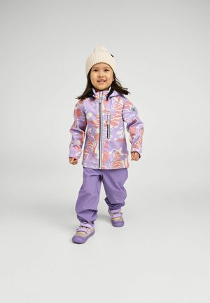 Niño pequeño con un gorro de punto crema, chaqueta de camuflaje púrpura y rosa, pantalones lavanda y zapatos a juego, caminando hacia adelante con una sonrisa.