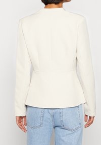 Blazer blanc sur mesure avec un ourlet à basque et des manches longues, présentant un design minimaliste et une texture lisse. Associé à un jean bleu clair.