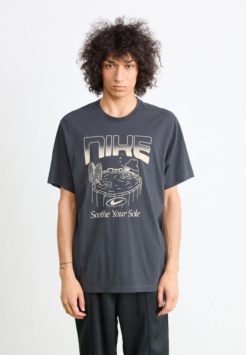 Ung person med krøllet hår iført en mørk Nike T-shirt med grafik af en sok, der ligger og svømmer i et badekar, med teksten "Soothe Your Sole."