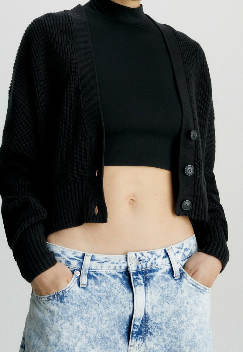 Černý žebrovaný kardigan s oversized rukávy, se třemi knoflíkovými uzávěry. V kombinaci s světlými modrými džínovými šortkami s vysokým pasem a roztrhaným efektem.