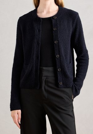 Femme portant un cardigan boutonné noir texturé, un haut côtelé noir et un pantalon taille haute noir, debout devant un fond uni.