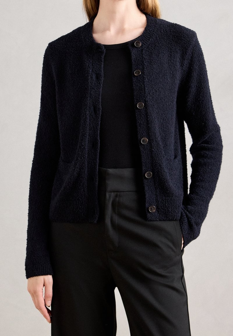 Femme portant un cardigan boutonné noir texturé, un haut côtelé noir et un pantalon taille haute noir, debout devant un fond uni.
