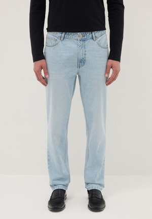 Man draagt lichtblauwe jeans met rechte pijpen, een zwart shirt met lange mouwen en zwarte loafers, staand tegen een effen achtergrond.