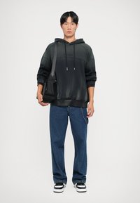 Fekete árnyalatú kapucnis pulcsi húzózsinórral, laza szabású, kék bő nadrággal és fekete-fehér tornacipővel párosítva. A modellt egy fekete táska tartja.