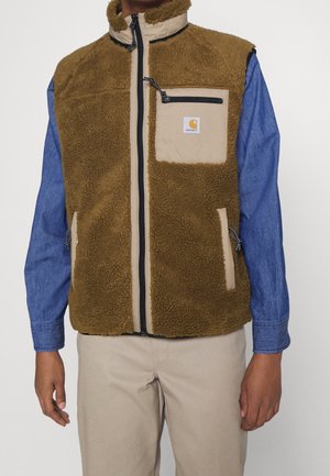 Homme portant un gilet polaire marron avec une bordure beige et une poche poitrine zippée, par-dessus une chemise bleue à manches longues et un pantalon beige.