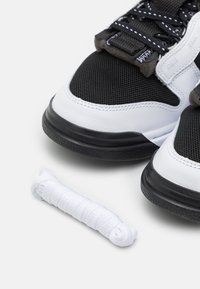 Sneakers in bianco e nero realizzati in materiali misti di rete e pelle, con suole testurizzate e un laccio bianco posizionato accanto.