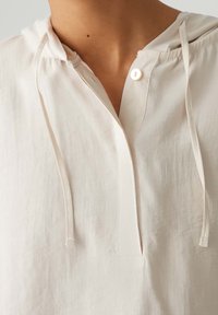 Jasno beżowy sweter z kapturem, plisą i zapięciem na guziki. Posiada miękką, teksturowaną tkaninę oraz sznurki przy szyi do regulacji.