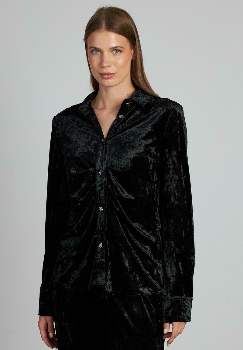 Camisa de botonadura negra de terciopelo con mangas largas, que presenta un corte holgado y un brillo sutil, incluye botones metálicos y detalles fruncidos.