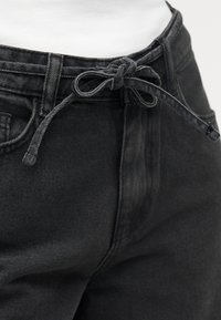 Κοντινή λήψη μαύρου τζιν παντελονιού με υφασμάτινη ζώνη δεμένη σε φιόγκο στη μέση, φορεμένο πάνω από λευκή μπλούζα.