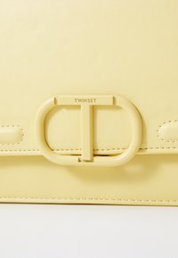 Yellow leather handbag with a smooth texture, featuring a prominent oval hardware closure and engraved "TWINSET" logo.