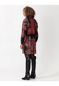 Schwarzes Kleid mit rotem Paisleymuster, langen Ärmeln und ausgestelltem Saum. Trägt kniehohe schwarze Lederstiefel, die einen Kontrast schaffen.