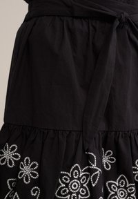 Zwart katoenen jurk met een aansluitende taille, een uitlopende rok en witte bloemige borduursels langs de zoom, voorzien van een touwstrikdetail in de taille.