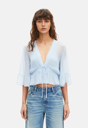 Kobieta ubrana w jasnoniebieską przezroczystą bluzkę z głębokim dekoltem w serek, z wiązaniem w talii i rozszerzanymi rękawami, w połączeniu z wyblakłymi niebieskimi dżinsami.