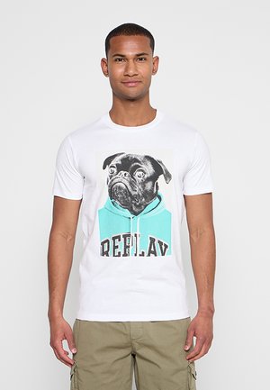 Mann trägt weißes T-Shirt mit schwarzem Mops, der eine türkise Kapuzenjacke und den Schriftzug „REPLAY“ trägt, kombiniert mit beigen Cargo-Shorts, steht vor schlichtem Hintergrund.