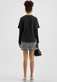 Czarny, oversize t-shirt nałożony na przezroczystą bluzkę z długim rękawem, zestawiony z mini spódnicą z srebnymi cekinami i małą czarną torebką.