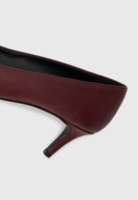 Scarpa con tacco alto in pelle bordeaux, con punta arrotondata, tacco sottile e texture liscia, caratterizzata da cuciture sottili lungo i bordi.