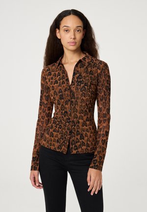 Een vrouw staat in een nauwsluitende bruine en zwarte luipaardprint blouse met knoopsluiting en zwarte broek voor een effen lichte achtergrond.