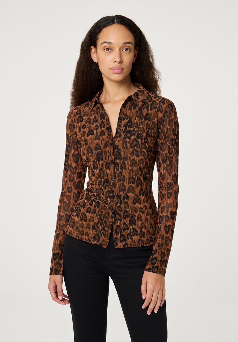 Een vrouw staat in een nauwsluitende bruine en zwarte luipaardprint blouse met knoopsluiting en zwarte broek voor een effen lichte achtergrond.