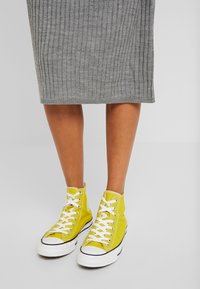 Des baskets montantes jaunes en toile présentent des lacets blancs et des semelles en caoutchouc. Le modèle porte une robe en tricot côtelé gris.
