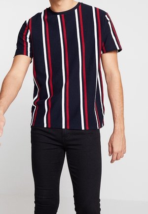 Mann, der ein kurzärmeliges marineblaues Hemd mit vertikalen roten, weißen und schwarzen Streifen sowie eine schwarze Hose vor einem schlichten Hintergrund trägt.