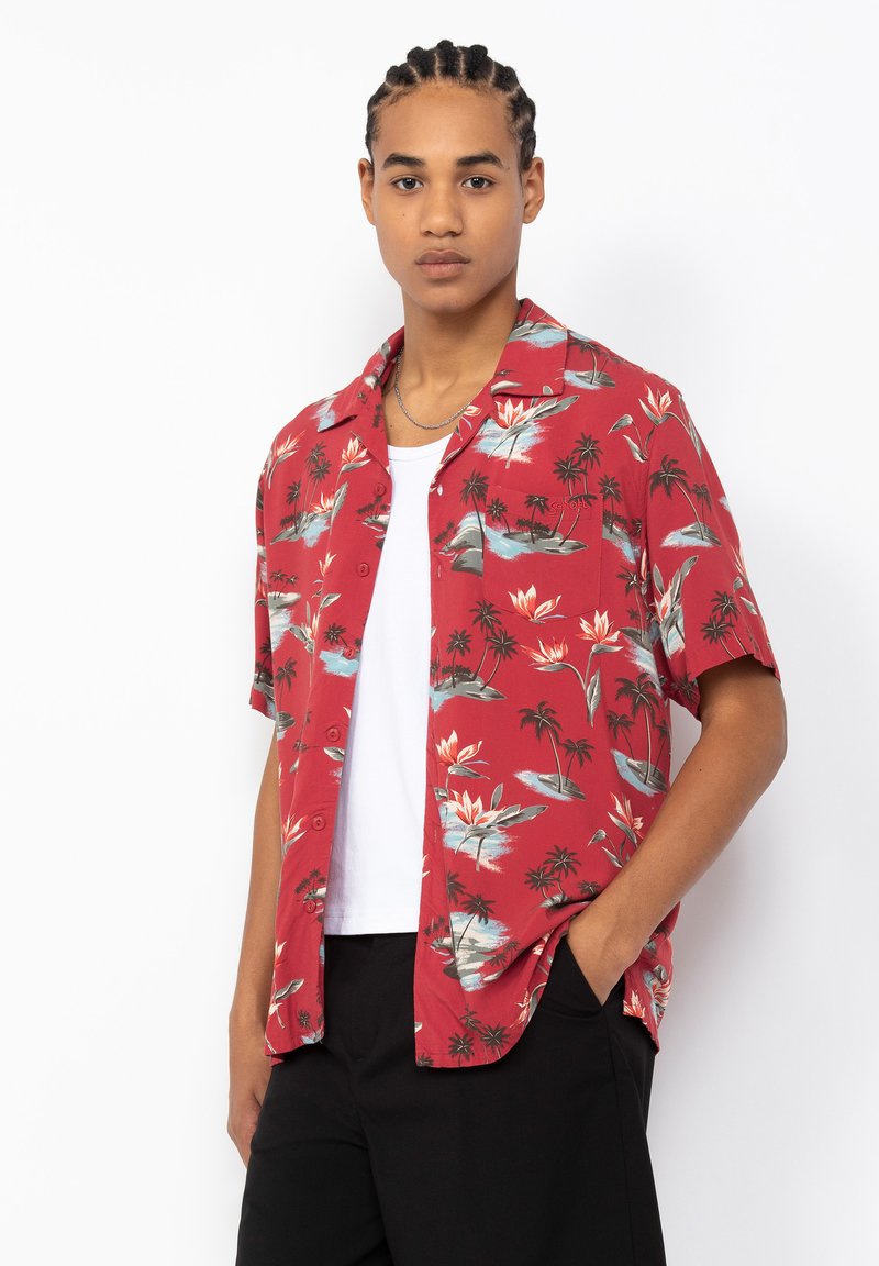 Rood overhemd met korte mouwen, gekenmerkt door een tropisch bloemen- en palmboompatroon. Met knoopsluiting aan de voorkant, kraag en een relaxte pasvorm. Gedragen over een wit T-shirt.