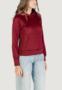 Felpa a cappuccio bordeaux con maniche lunghe, dotata di tasca a marsupio e orlo aderente. Tessuto liscio, materiale elastico e branding minimal.