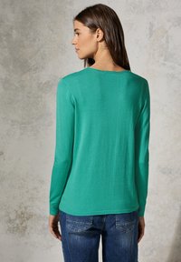 Grünes Langarmtop aus weichem Material mit rundem Halsausschnitt, das eine glatte Textur und eine figurbetonte Silhouette aufweist, kombiniert mit blauen Jeans.