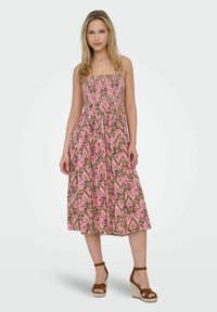 Lång, rosa mönstrad klänning med en figursydd överdel, smockdetaljer och en flödande kjol, kombinerad med bruna kilklackssandaler.