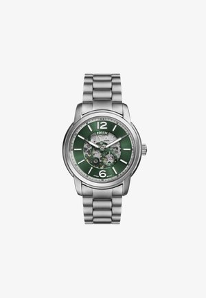 Montre en acier inoxydable argenté avec un cadran vert, un boîtier transparent, des index horaires blancs et un bracelet en maille. Elle présente des mécanismes internes visibles.
