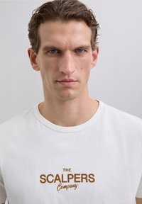 Uomo con capelli corti e ricci di colore mogano che indossa una maglietta bianca con il testo "THE SCALPERS Company" in marrone su uno sfondo chiaro e semplice.
