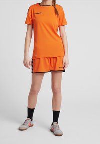 Camiseta y pantalones cortos deportivos de un brillante color naranja con acentos negros, que presentan el logo de Hummel. El modelo lleva zapatillas deportivas grises y rojas con calcetines negros.