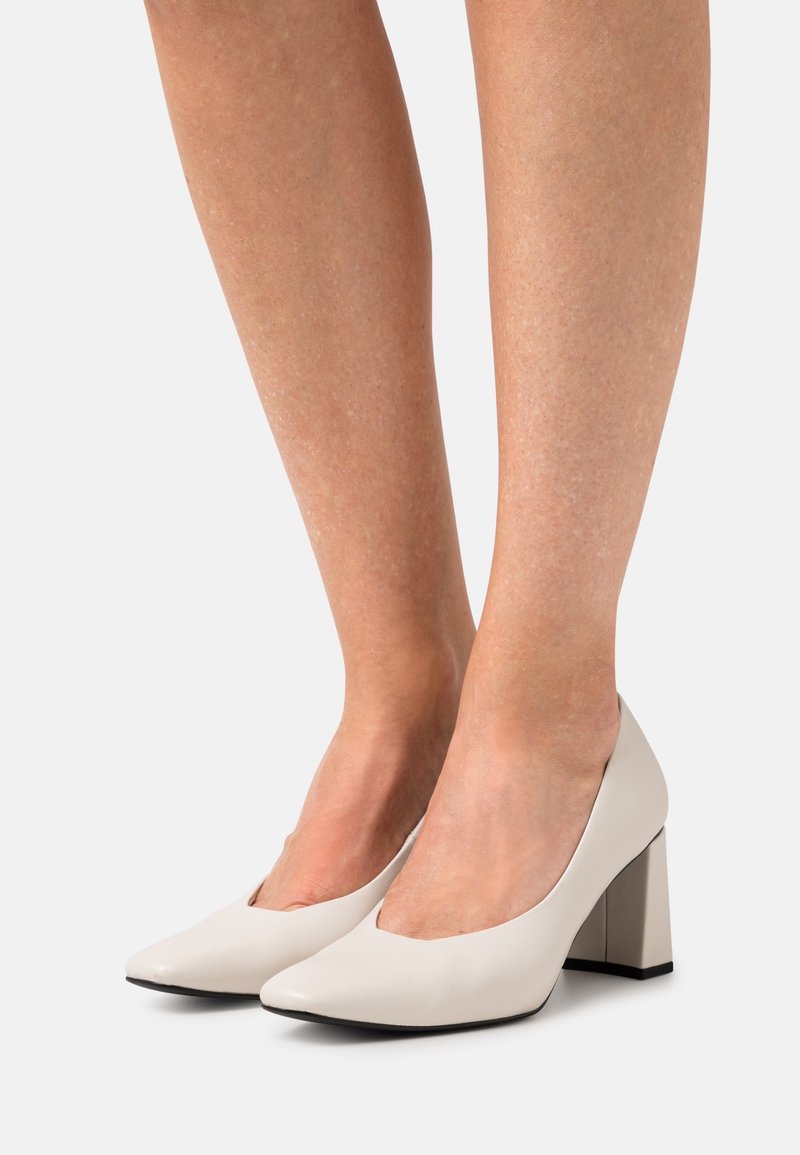 Jambes portant des chaussures blanches à talon bloc à bout pointu avec une finition lisse sur un fond clair uni.