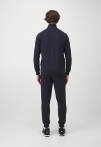 Marinträningsoverall med en zip-up jacka och elastiska muddar, med en slät yta. Svarta träningsskor fulländar outfiten.