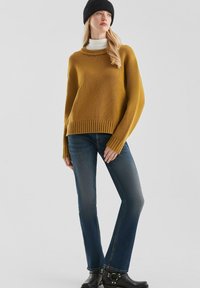 Mosterdkleurige gebreide trui met geribbelde zoom, gecombineerd met blauwe jeans. Zwarte beanie en enkellaarsjes met zilverkleurige hardware.