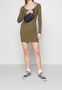 Kvinna klädd i långärmad mini-klänning i olivgrön, som håller en svart axelväska, med vita strumpor och olivgröna sneakers.