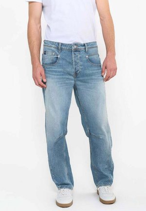 Jean en denim bleu clair délavé avec une coupe relaxed, cinq poches et de subtils détails de couture, porté avec un t-shirt blanc et des baskets blanches.