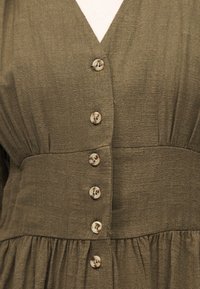 Φόρεμα από ύφασμα ελιάς σε πράσινο χρώμα με V-λαιμόκοψη, σφηκοειδή μέση και σειρά από έξι στρογγυλά, μπεζ κουμπιά μπροστά.