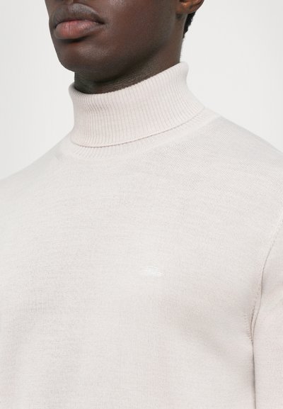 Primo piano di una persona che indossa un maglione a collo alto a coste di un beige chiaro, con un logo ricamato discreto sul petto.