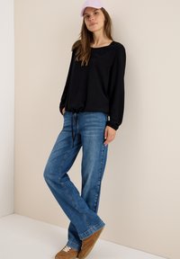 Zwarte longsleeve top met een trekkoord aan de onderkant, gecombineerd met lichtblauwe wijde jeans. Model draagt een lichtpaarse pet en bruine schoenen.