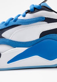 Gros plan d'une basket de sport bleue, blanche et noire montrant le tissu en mesh, les surcouches en daim, les lacets et les détails de la semelle coussinée.
